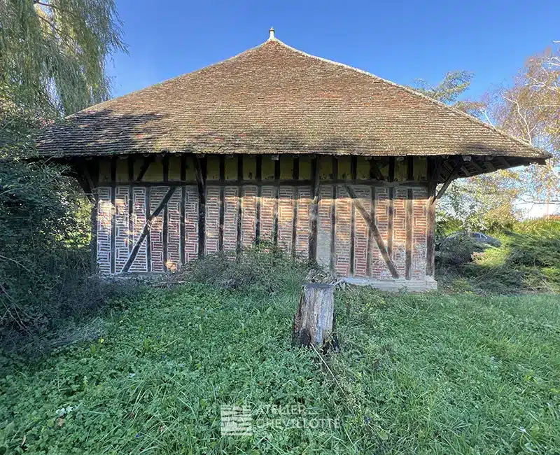 Bâtiment ancien en colombages avec remplissage en briques, large toiture à quatre pans et façade conservée, en attente de restauration.