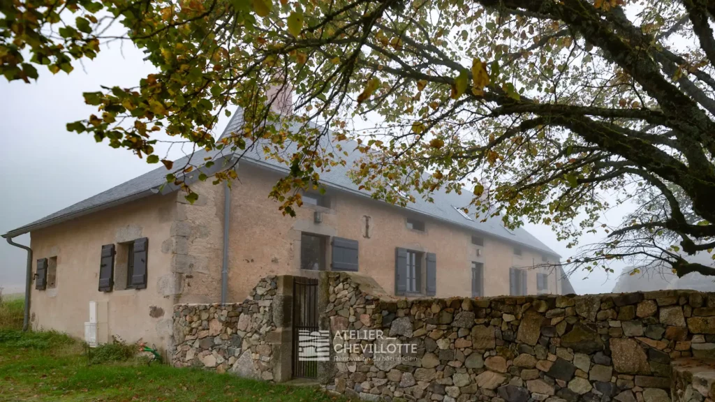 Maison paysanne traditionnelle en pierre du Morvan, après réhabilitation par l’Atelier Chevillotte, entourée d’un mur en pierre sèche et d’un grand arbre, dans une ambiance brumeuse et naturelle.